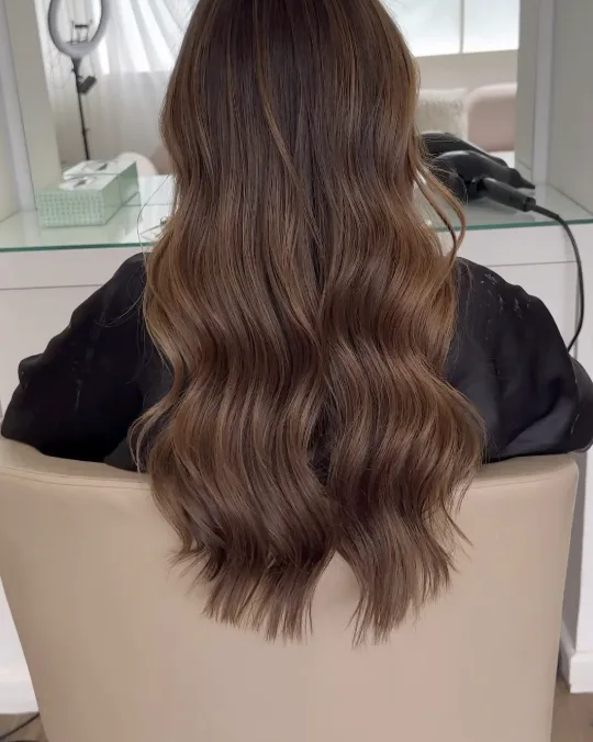 Balayage brun ondulé vu de dos, devant le miroir du salon