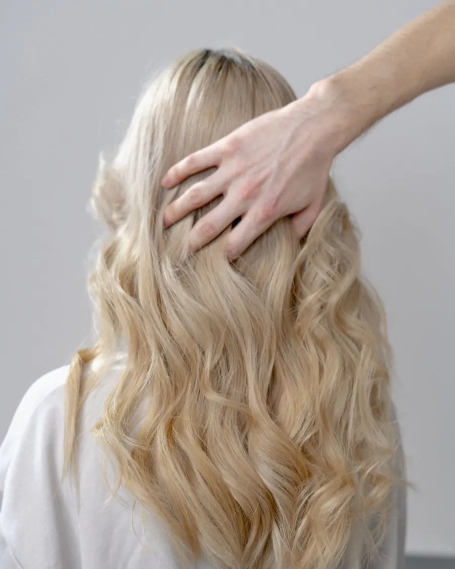 Longue chevelure blonde bouclée, vue de dos, travaillée par la main d'une coiffeuse
